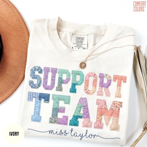 May include: Ivory-colored t-shirt with the words "SUPPORT TEAM" in patchwork letters. The letters are in various pastel colors and patterns. The name "miss taylor" is written in cursive below the words. A gold necklace and a brown hat are also visible.