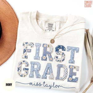 May include: Ivory-colored t-shirt with the words "FIRST GRADE" in blue patchwork letters. The letters are decorated with floral, polka dot, and gingham patterns. The name "miss taylor" is written in cursive below the text. A gold necklace is draped across the shirt.
