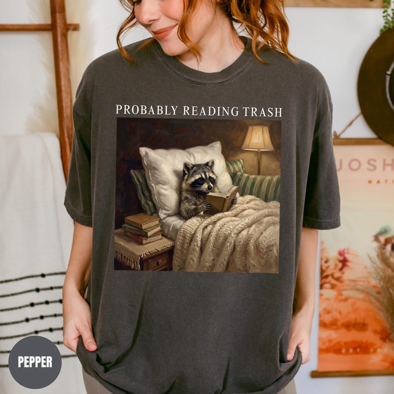 以下が含まれることがあります： 「PROBABLY READING TRASH」の文字がプリントされた濃いグレーのTシャツ。ベッドで本を読むアライグマの画像が上にあります。アライグマは枕とランプのある毛布の下にいます。