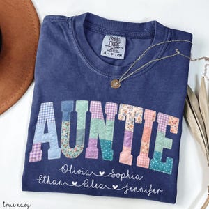 May include: A navy blue t-shirt with the word "AUNTIE" in patchwork letters. The names Olivia, Ethan, Sophia, Alex, and Jennifer are printed below the word. A gold necklace with a pendant is draped across the shirt.