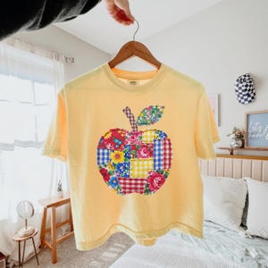 May include: A pale yellow short-sleeve t-shirt featuring a large central apple graphic composed of various colorful patchwork fabric squares, including floral, gingham, and plaid patterns in red, blue, yellow, and green, hanging on a wooden hanger.