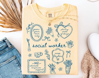 May include: A yellow "Comfort Colors" sweatshirt with blue illustrations and text. The design includes floral elements, bows, and the words "social worker." Additional text includes "grow your own way," "planting seeds of change," and "advocate empower support."