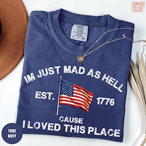 May include: A navy blue t-shirt with the text "IM JUST MAD AS HELL CAUSE I LOVED THIS PLACE" and an American flag graphic. The shirt also has the text "EST. 1776". The shirt is folded and has a Comfort Colors tag.