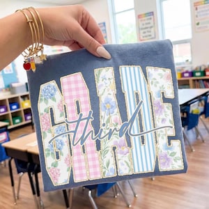 May include: A slate blue t-shirt featuring the word GRADE in large block letters with floral and pink gingham patterns, overlaid with the word third in cursive script. The shirt is held up, displaying gold-toned bangles with school-themed charms.
