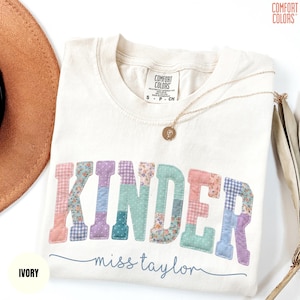 May include: Ivory-colored Comfort Colors t-shirt with the word "KINDER" in patchwork letters. The letters are in pastel colors and floral patterns. The name "miss taylor" is written in a script font below the word "KINDER". A gold necklace is draped across the shirt.