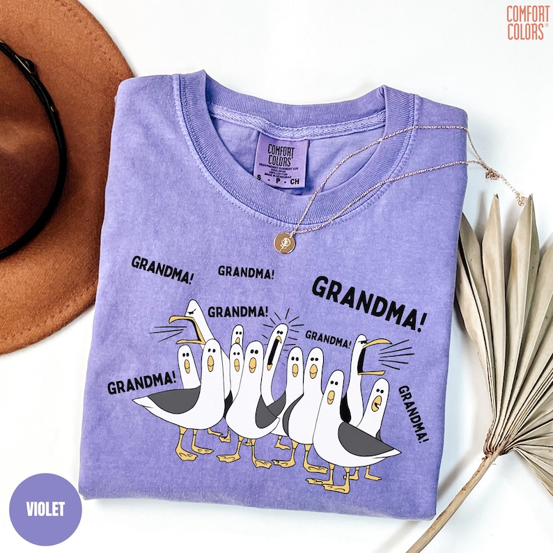 May include: A violet Comfort Colors sweatshirt with a graphic of seagulls and the word "GRANDMA!" repeated around them. The shirt has a crew neck and long sleeves. A gold necklace and a brown hat are also in the image.