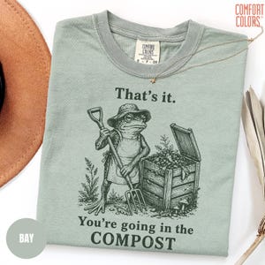 May include: A light green t-shirt with the text "That's it. You're going in the COMPOST" and a frog graphic. The frog is wearing a hat and apron, holding a shovel and pitchfork. The shirt is a Comfort Colors brand.