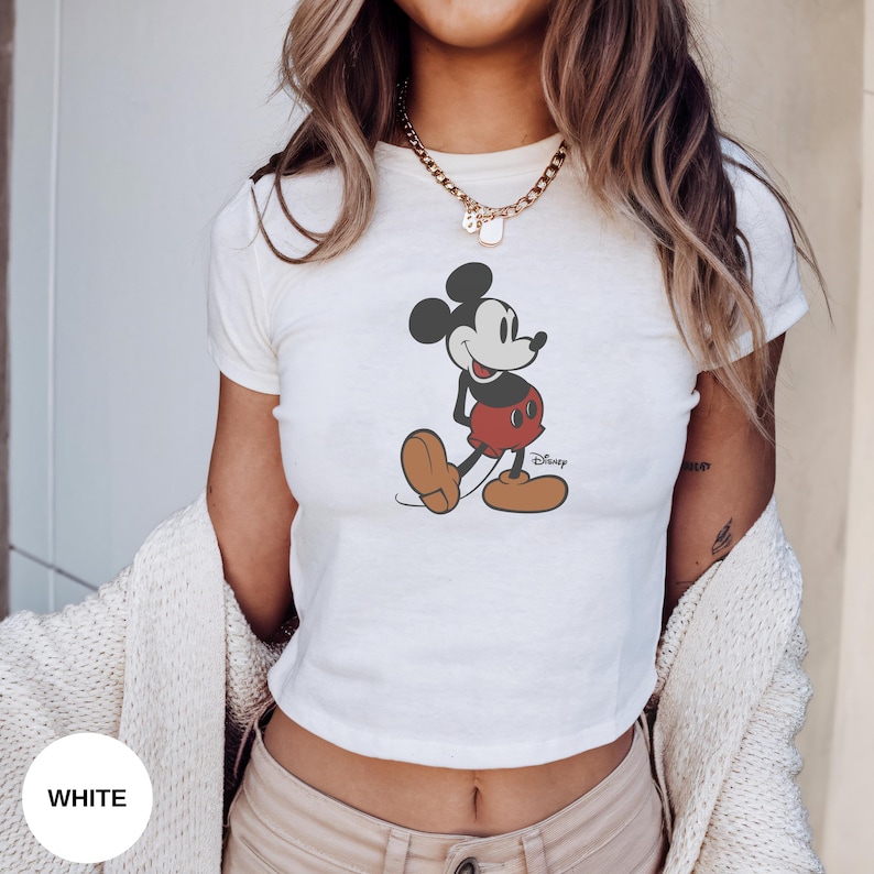 Puede incluir: Top corto blanco con un gr&aacute;fico de Mickey Mouse. El personaje de dibujos animados est&aacute; representado en negro, rojo y marr&oacute;n. El logotipo de Disney es visible. El top es de manga corta.