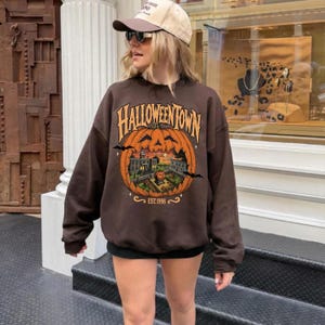 May include: A dark brown crewneck sweatshirt featuring a large graphic of a carved pumpkin with a house scene inside. The text reads Halloweentown Est. 1998 in a stylized orange font. The design includes small black bats and stars on the pumpkin.