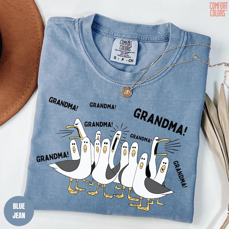 May include: A blue jean Comfort Colors t-shirt with a graphic of seagulls and the word "GRANDMA!" repeated around them. The shirt has a crew neck and short sleeves. A gold necklace with a pendant is draped across the shirt.