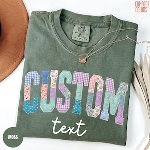 May include: A moss green t-shirt with the word "CUSTOM" in patchwork letters. The letters are made of various floral and patterned fabrics. The word "text" is written below in a cursive font. A gold necklace rests on the shirt.