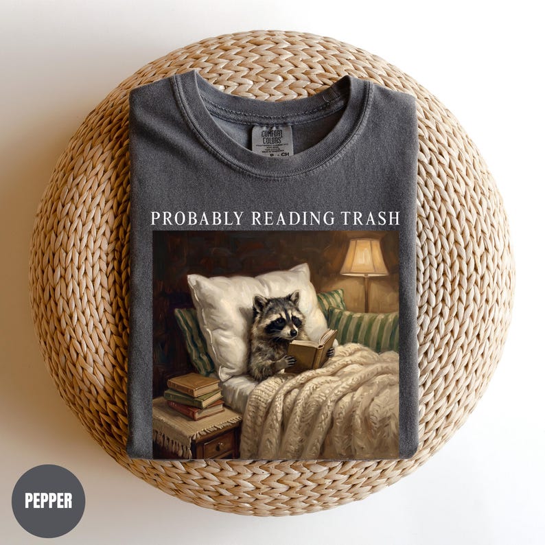 以下が含まれることがあります： 「PROBABLY READING TRASH」の文字が入った濃いグレーのTシャツ。 ランプと枕のある居心地の良い環境で本を読むアライグマが描かれています。 Tシャツは織られた表面に折りたたまれています。