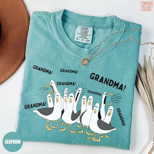 May include: Seafoam green t-shirt with a graphic of seagulls yelling "GRANDMA!". The shirt has a crew neck and short sleeves. A gold necklace with a pendant is draped across the shirt. The shirt is labeled "COMFORT COLORS".