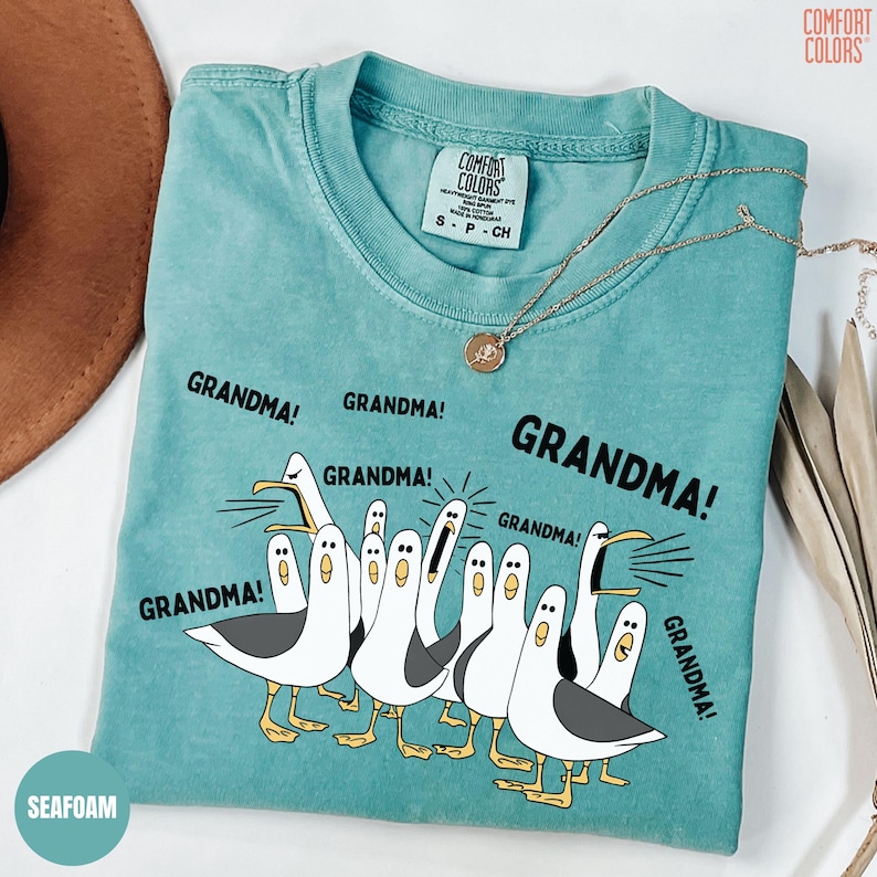 May include: Seafoam green t-shirt with a graphic of seagulls yelling "GRANDMA!". The shirt has a crew neck and short sleeves. A gold necklace with a pendant is draped across the shirt. The shirt is labeled "COMFORT COLORS".