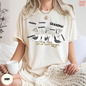 May include: Ivory-colored t-shirt featuring a cartoon of seagulls with the word "GRANDMA!" above each bird. The Comfort Colors brand logo is visible in the upper right corner. The shirt is worn by a person.