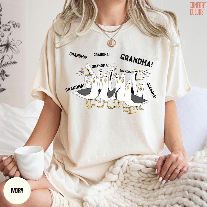 May include: Ivory-colored t-shirt featuring a cartoon of seagulls with the word "GRANDMA!" above each bird. The Comfort Colors brand logo is visible in the upper right corner. The shirt is worn by a person.