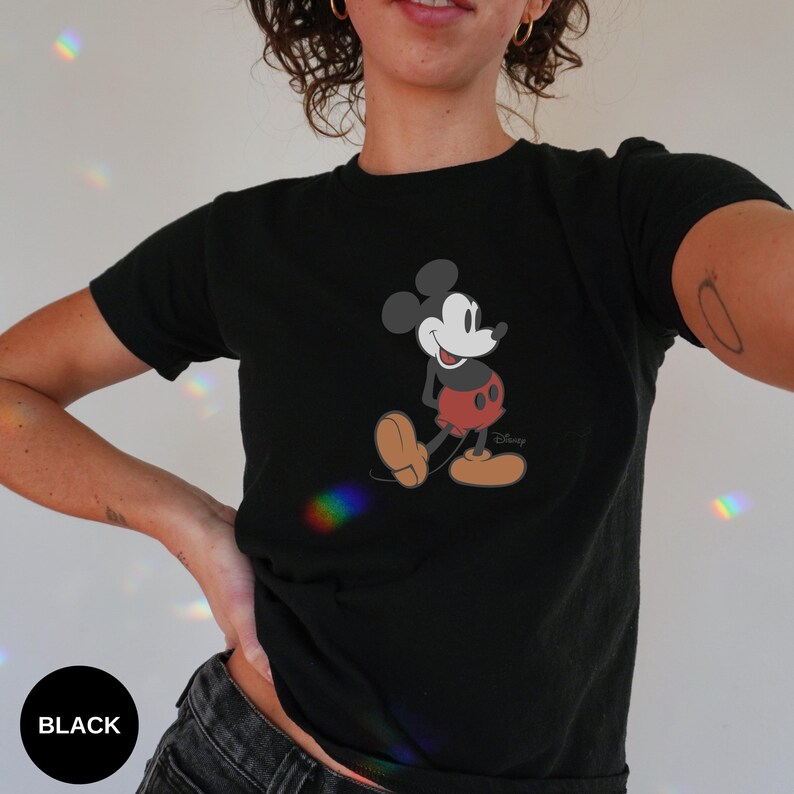 Puede incluir: Camiseta negra con un dibujo de Mickey Mouse caminando. El personaje de dibujos animados est&aacute; representado en sus colores cl&aacute;sicos: negro, rojo, beige y blanco. El logotipo de Disney es visible en la parte inferior derecha.