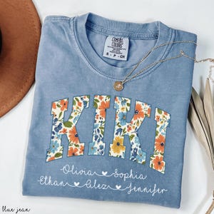 May include: A blue Comfort Colors t-shirt with the name "KIKI" in floral print letters. Below the name are the names Olivia, Sophia, Ethan, Alex, and Jennifer. A gold necklace with a pendant rests on the shirt.
