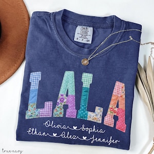 May include: A navy blue Comfort Colors t-shirt with the word "LALA" in patchwork letters. Below the letters are names and hearts. A gold necklace rests on the shirt. The shirt is labeled "true navy".