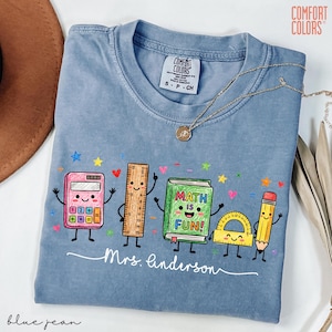 May include: A blue crew neck t-shirt featuring a graphic of cartoon school supplies including a pink calculator, a wooden ruler, a green book with the text "MATH IS FUN!", a yellow protractor, and a yellow pencil, with the name "Mrs. Anderson" below.