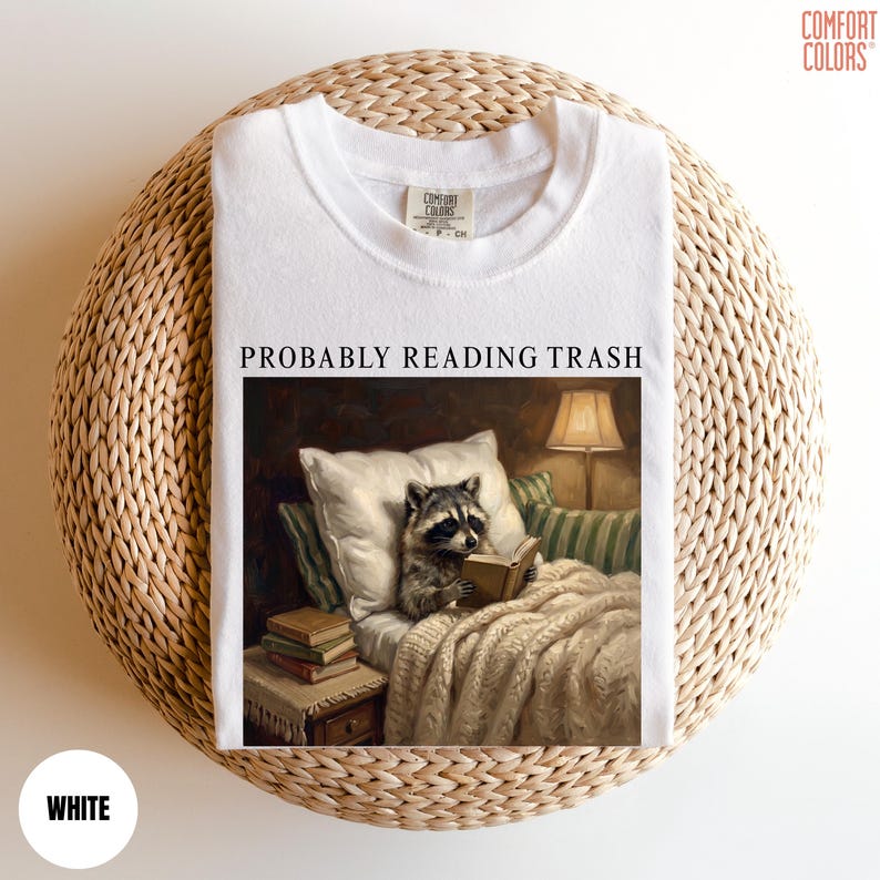 以下が含まれることがあります： 「PROBABLY READING TRASH」の文字と、ベッドで本を読むアライグマのイラストがプリントされた白いTシャツ。アライグマはランプと枕のある居心地の良い環境にいます。このシャツは柔らかい素材で作られています。