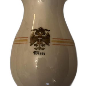 May include: Cream-colored ceramic vase with a flared top and rounded body. It features a gold and brown emblem with the word "Wien" beneath it. Two gold stripes encircle the vase.