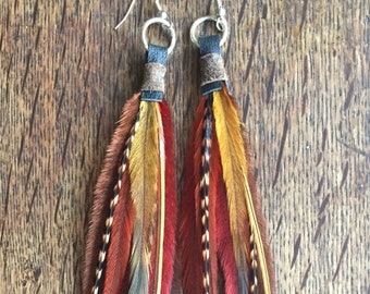 May include: A pair of long feather earrings with brown leather cord accents. The feathers are red, orange, yellow, and black.