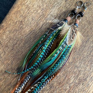 Puede incluir: Un par de pendientes largos de plumas con una variedad de colores, incluyendo verde, turquesa, marrón y naranja. Los pendientes tienen aros plateados y están adornados con cuentas y detalles de cuero. Las plumas están dispuestas en un diseño en capas.
