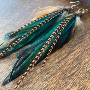May include: A pair of long, dangling earrings featuring teal and brown feathers. The feathers are arranged in a layered pattern, with the teal feathers on top and the brown feathers underneath. The earrings are attached to silver hooks.