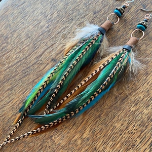 Puede incluir: Un par de pendientes largos y colgantes con cordón de cuero marrón, cuentas turquesa y negras y múltiples plumas en tonos marrones, verdes y azules.