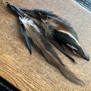 May include: A handcrafted feather earring featuring dark brown and black feathers, accented with leather straps and gold-toned metal details. The earring has a bohemian style, with a natural and earthy aesthetic.
