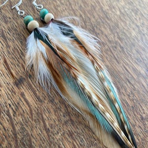 May include: A pair of feather earrings with silver hooks, wooden beads, and teal beads. The feathers are a mix of white, brown, teal, and black. The earrings are handmade.