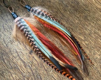 May include: A pair of long dangle earrings featuring layered feathers in shades of burnt orange, turquoise, brown, and black. Each earring is attached to a bronze-toned metal ear wire with a small black leather wrap securing the feather cluster.