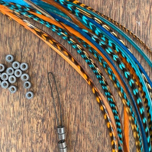 May include: A selection of vibrant hair feathers in blue, orange, and brown, alongside small metal beads and a loop tool for application. The items are displayed on a wooden surface.