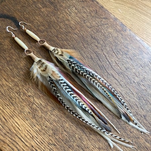 May include: A pair of long feather earrings with a bone bead accent. The feathers are white, brown, black, and blue.