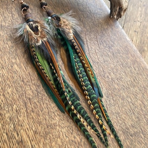May include: Long feather earrings with a mix of green, brown, and orange feathers. The earrings have a gold-colored hook and a dark brown leather accent. The feathers are arranged in a layered design, creating a unique and eye-catching accessory.