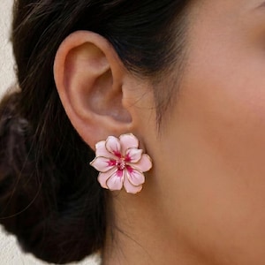 May include: Pink flower-shaped stud earrings with gold-colored trim. The earrings have a detailed design with a darker pink center and lighter pink petals. The earrings are worn on an ear.