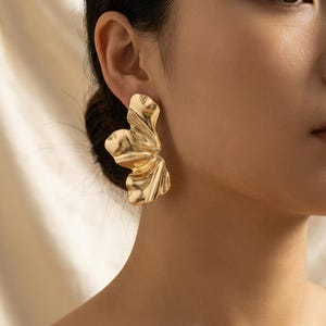 May include: Gold-colored statement earrings with a textured, abstract floral design. The earrings have a layered, petal-like appearance, reflecting light and adding a touch of glamour. These earrings are a bold accessory for any outfit.