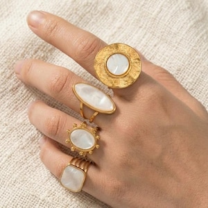 May include: A hand displays four gold-colored rings. Each ring features a different design, including a large circular ring with a white center, an oval ring, a ring with a sunburst design, and a square ring.