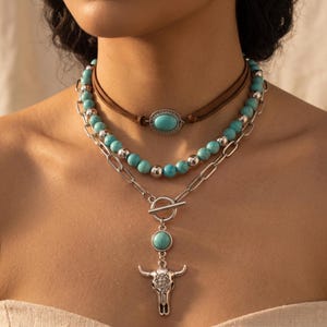 May include: A layered necklace set featuring a brown cord choker with an oval turquoise stone, a strand of round turquoise and silver beads, and a silver chain with a toggle clasp and a silver bull skull pendant with a turquoise stone.