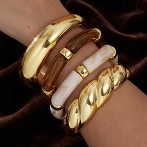May include: A selection of bracelets in gold and neutral tones. The bracelets include a thick, solid gold bangle, a brown and gold beaded bracelet, a white and gold marbled bracelet, and a gold twisted design bracelet. The bracelets are worn on a wrist.