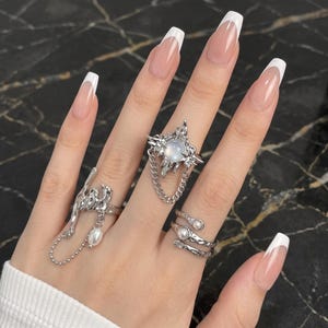 May include: Three silver-toned rings worn on fingers. One ring features a central iridescent orb with a dangling chain. Another ring has an abstract, fluid metal design with a small hanging pearl. The third ring is a wrap-style band with two pearls.