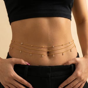 May include: Gold-colored body chain with three strands. The top strand has small gold beads, the middle strand has small clear gems, and the bottom strand has dangling clear gems. The body chain is worn around the waist.