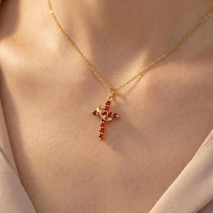 May include: A gold-colored necklace with a cross pendant. The cross is adorned with red gemstones and features a crown design at the top. The necklace is displayed against a neutral background.