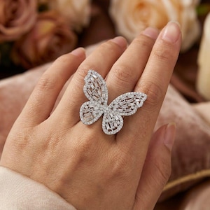 Könnte beinhalten: Ein silberfarbener Schmetterlingsring mit einem großen, komplizierten Design, das mit zahlreichen kleinen, klaren Baguette- und Rundschliffsteinen besetzt ist. Die Schmetterlingsflügel sind mit einer Pavé-Fassung versehen, die eine funkelnde Oberfläche erzeugt.