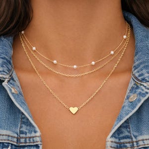 May include: A multi-strand gold necklace. The top two chains have small pearls, while the lowest has a heart pendant. The necklaces are shown against a denim jacket.