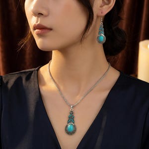 May include: A silver-coloured jewellery set comprising a necklace and earrings. The necklace features a pendant with a large oval turquoise stone, framed decoratively. The earrings echo the pendant design. The jewellery is worn against a dark blue top.