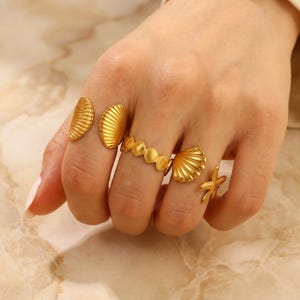 Puede incluir: Una mano con cinco anillos dorados. Los anillos presentan diseños de conchas marinas y estrellas de mar. La mano es de tono claro, sobre un fondo neutro.