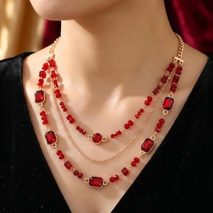 May include: A three-strand necklace featuring red rectangular and round gemstones, and small red beads. The necklace has a gold-colored chain and clasp. The jewelry is displayed against a black background.