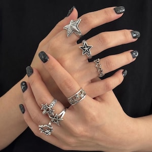May include: A collection of silver-toned metal rings worn on multiple fingers. The rings feature various star-shaped designs, including solid stars, open-work star bands, and pentagram motifs, all with an oxidised finish to highlight the details.